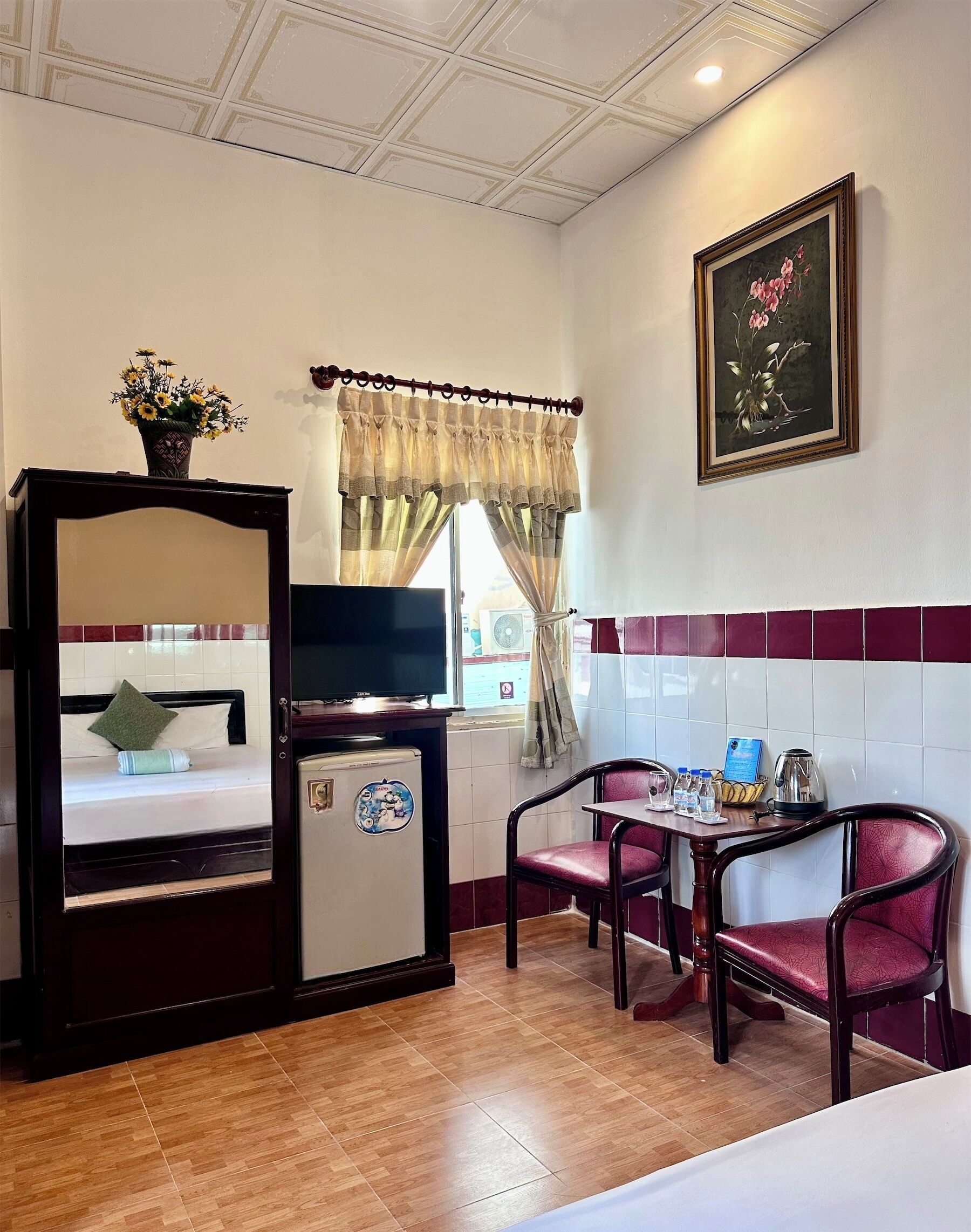 A hotel room with a bed, a TV, a small refrigerator, a dining table with chairs, and a window with curtains.