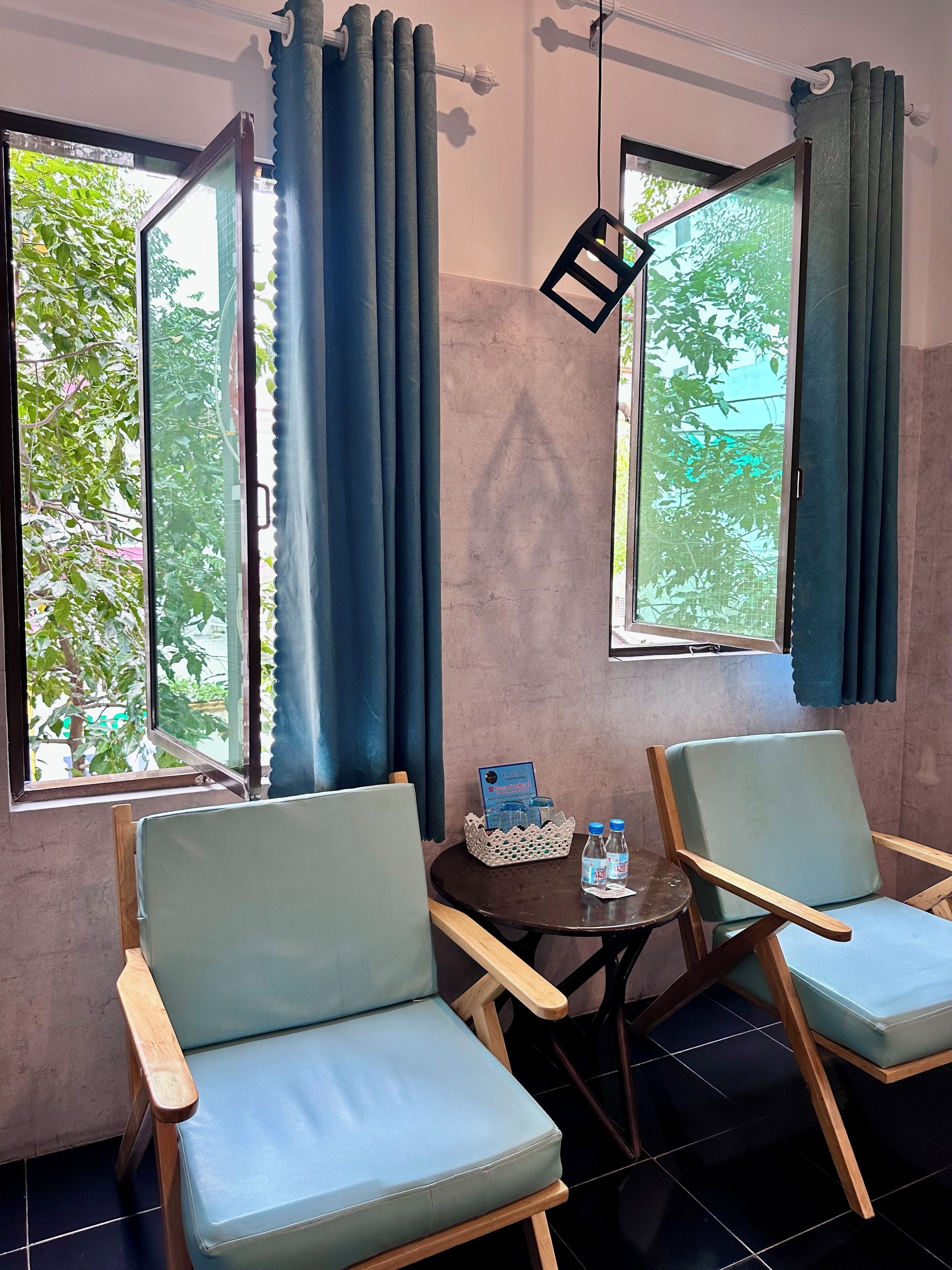 Una habitación con dos sillas azul claro, una mesita con una cesta que contiene botellas de agua y un cartel, y vistas a zonas verdes a través de las ventanas.
