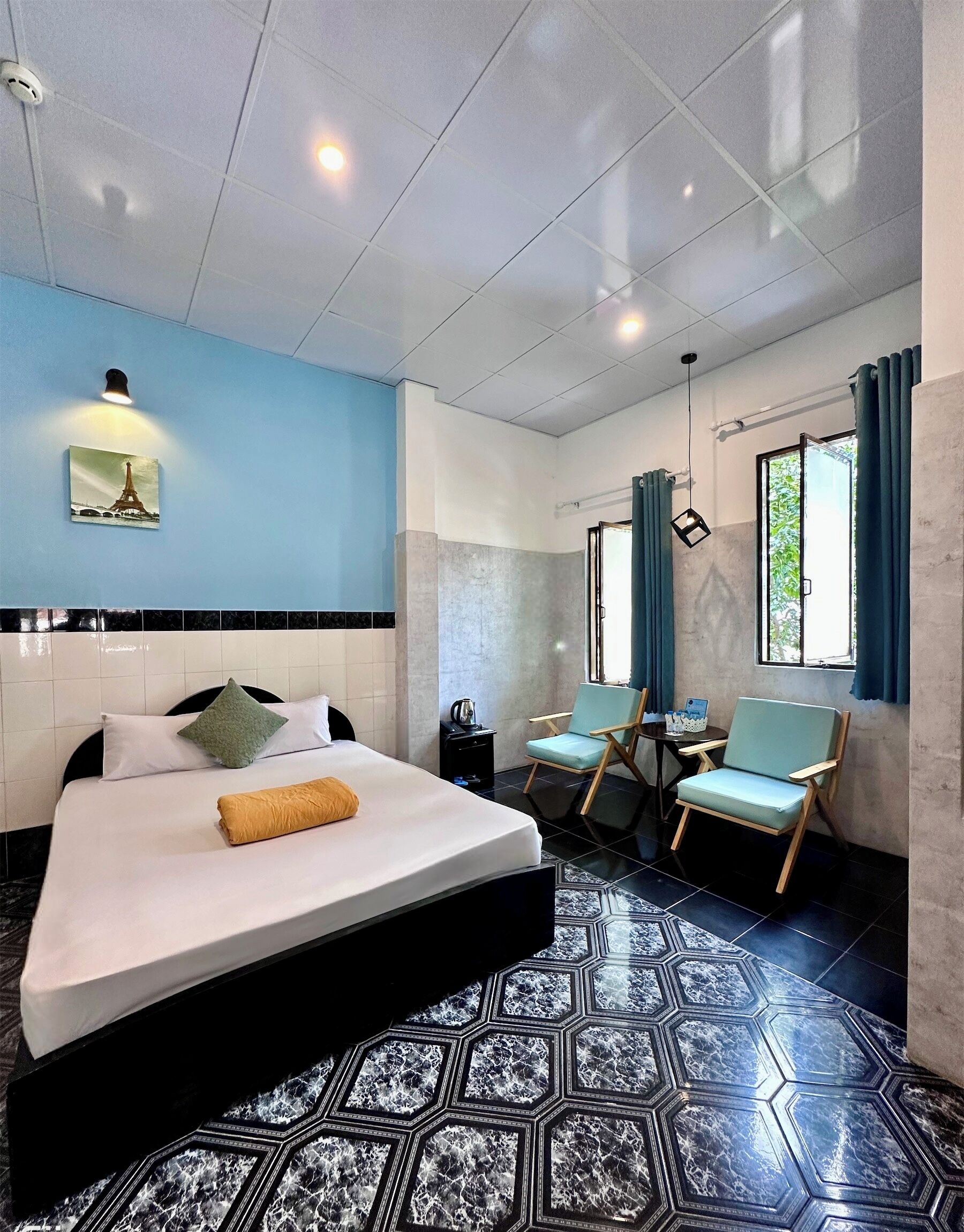 A hotel room with a bed, two chairs, a small table, a window with curtains, and a wall-mounted picture.