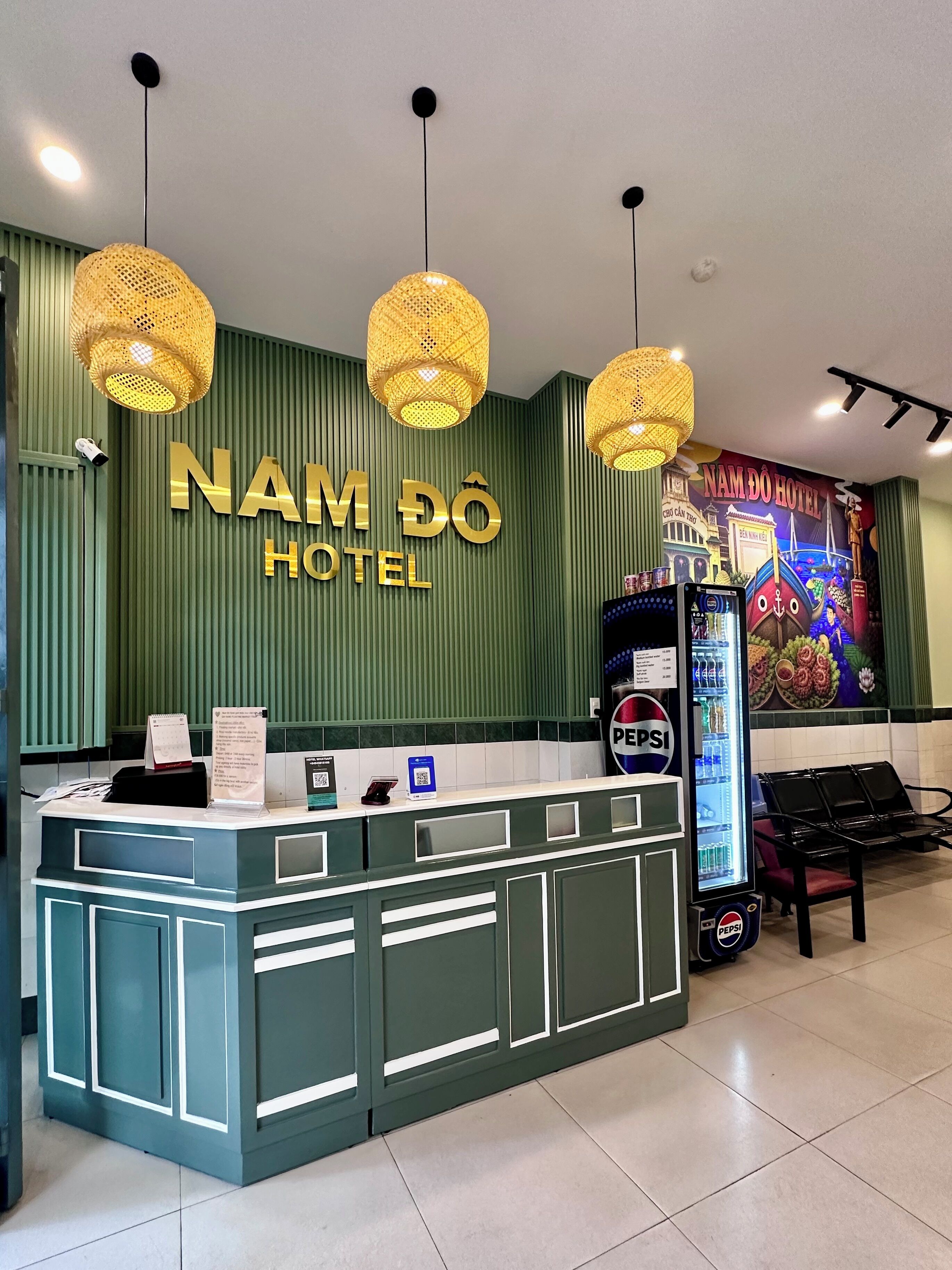 A reception desk with a wall sign for Nam Do Hotel, a security camera, a digital screen, and a Pepsi refrigerator.