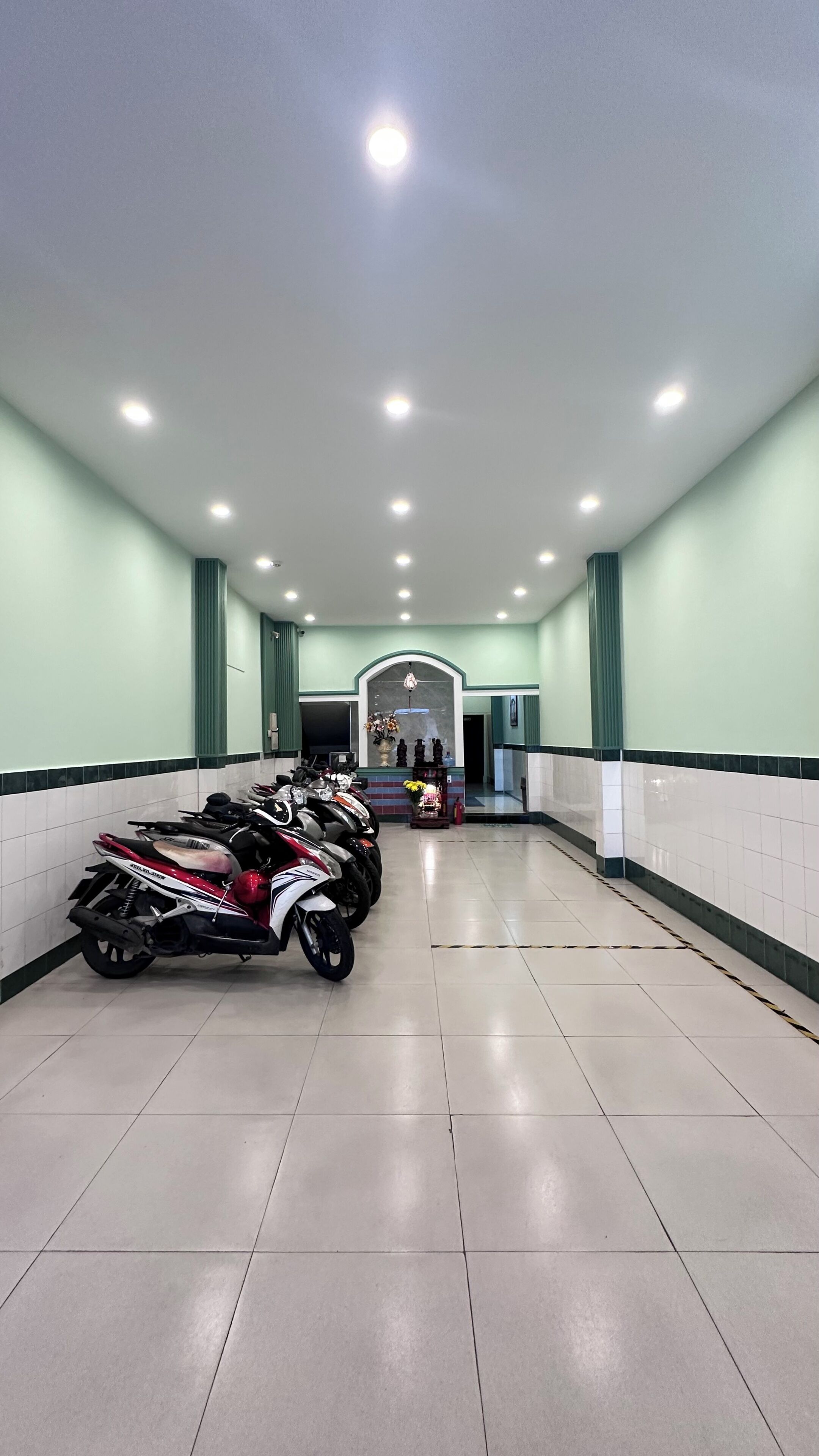 A clean, well-lit indoor space with a row of parked scooters, a tiled floor, and a wall-mounted display case with flowers.