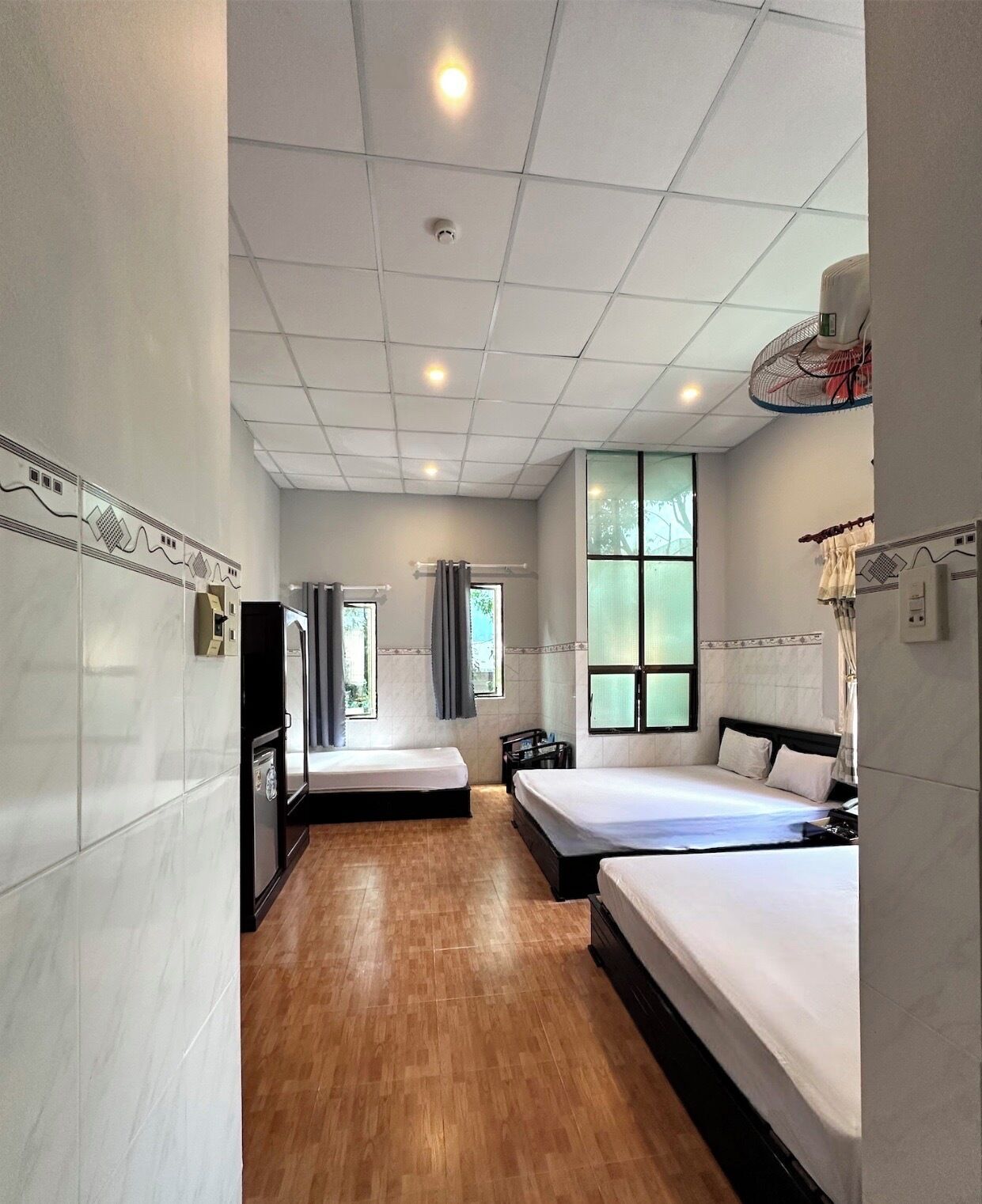A hotel room with two beds, a small kitchenette, a ceiling fan, and a window with curtains.