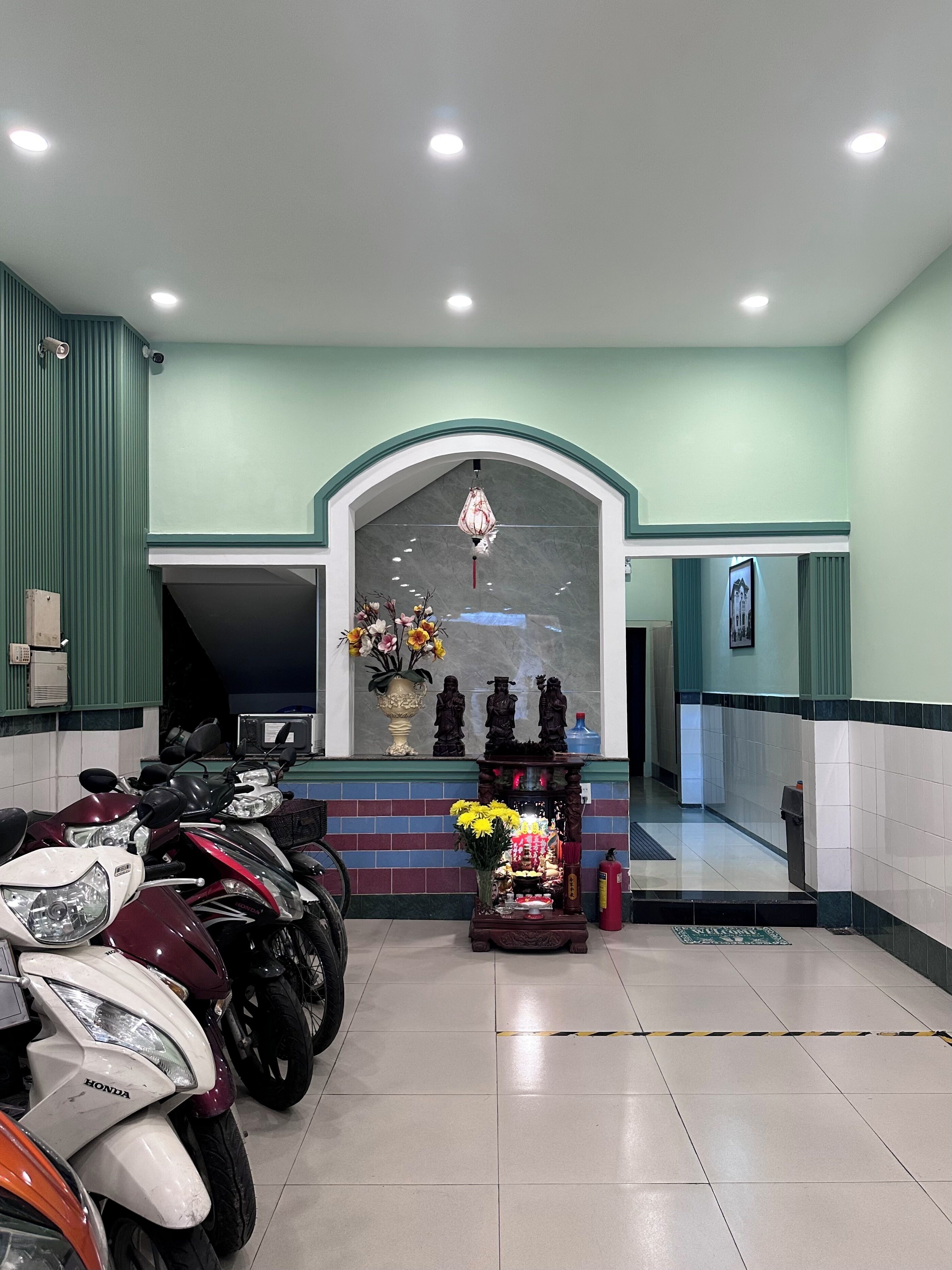 A room with a tiled floor, a green wall, and a ceiling with recessed lighting. There are several motorcycles parked in the room.