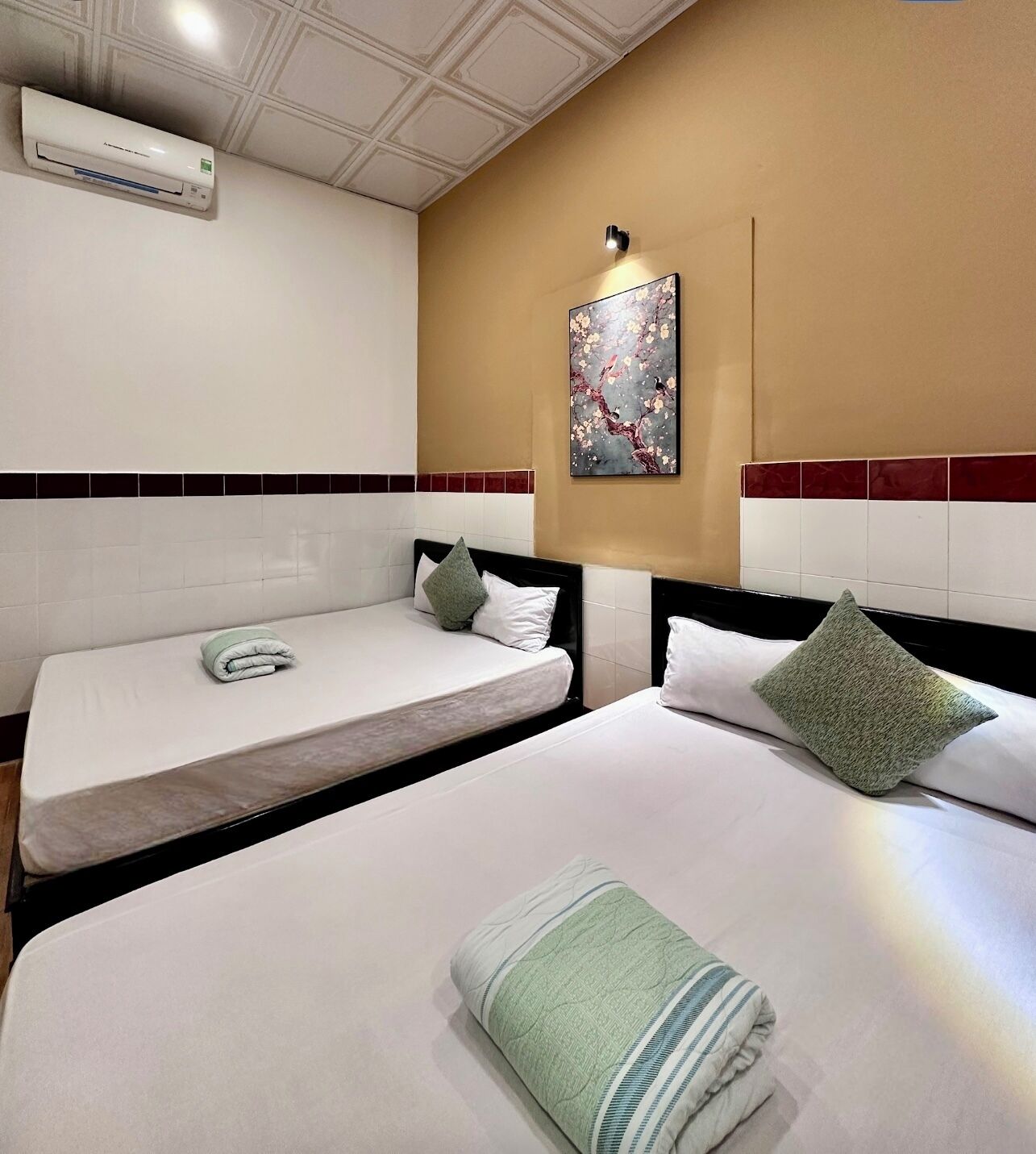 A hotel room with two beds, a wall-mounted air conditioner, a decorative painting, and a tiled backsplash.