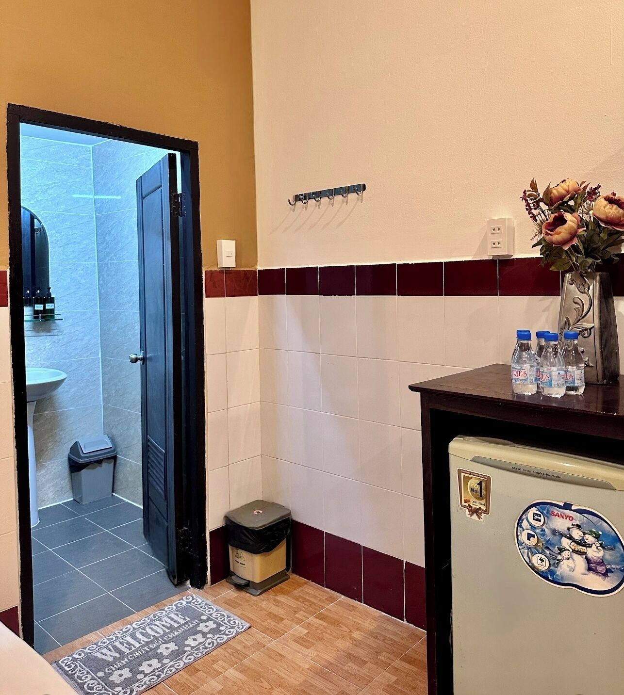Un baño de hotel con suelo de baldosas, un lavamanos, un cubo de basura y un pequeño refrigerador con un adhesivo de Sanyo.