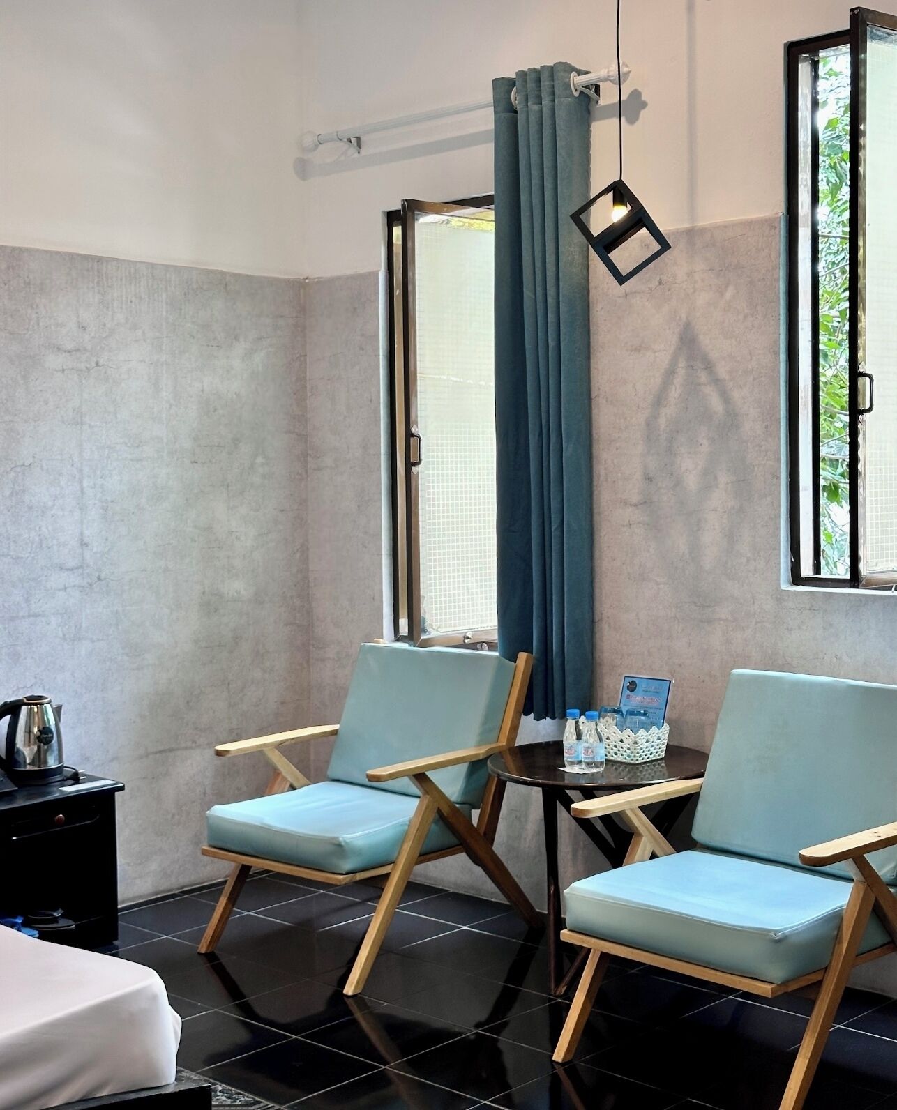 Una habitación con dos sillas azul claro, una mesita con un libro y botellas de agua, un hervidor negro en un mueble, y una ventana con cortina.
