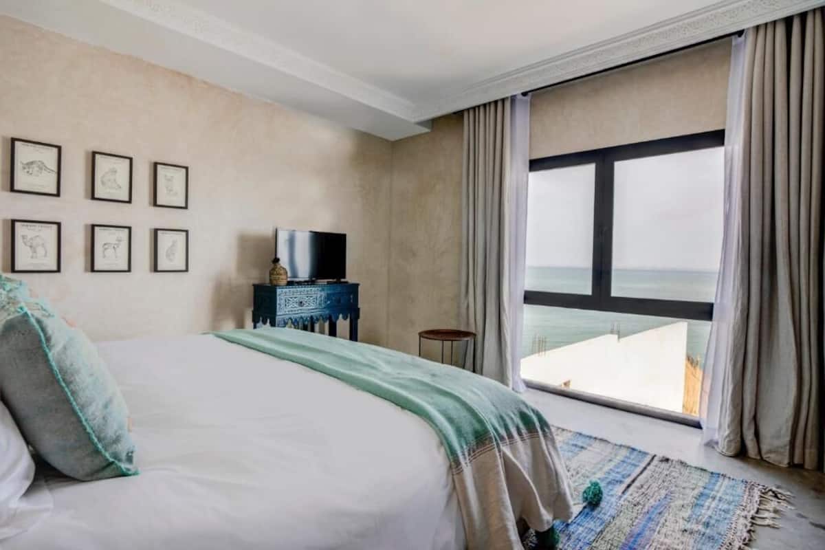 A hotel room with a large bed, a television on a side table, framed pictures on the wall, and a view of the sea through a large window.