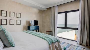 A hotel room with a large bed, a television on a side table, framed pictures on the wall, and a view of the sea through a large window.