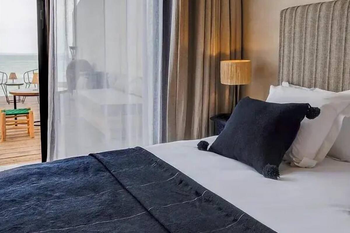 A hotel room with a bed, a nightstand, a lamp, and a view of the sea through large windows.