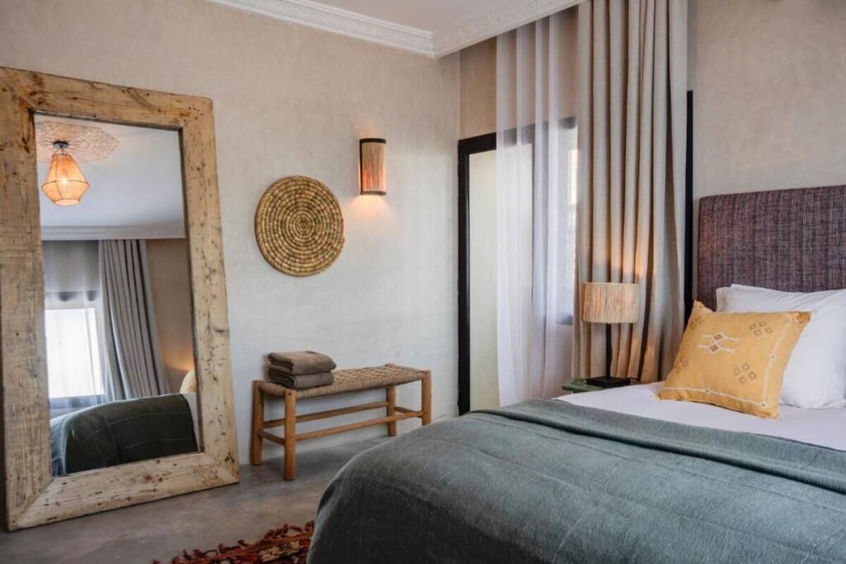 A hotel room with a large mirror, a bedside lamp, a bed with a green bedspread, a small wooden bench, and a wall-mounted woven art piece.