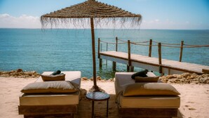 Un espacio de descanso frente al mar con dos sillones acolchados, una mesita redonda y un toldo de paja. Un muelle de madera se adentra en el agua.