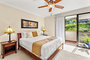 A hotel room with a large bed, two bedside lamps, a ceiling fan, a painting on the wall, and a view of a garden through a sliding door. - Kapalua Golf Villa, fresh updates and fairway view (Lahaina)