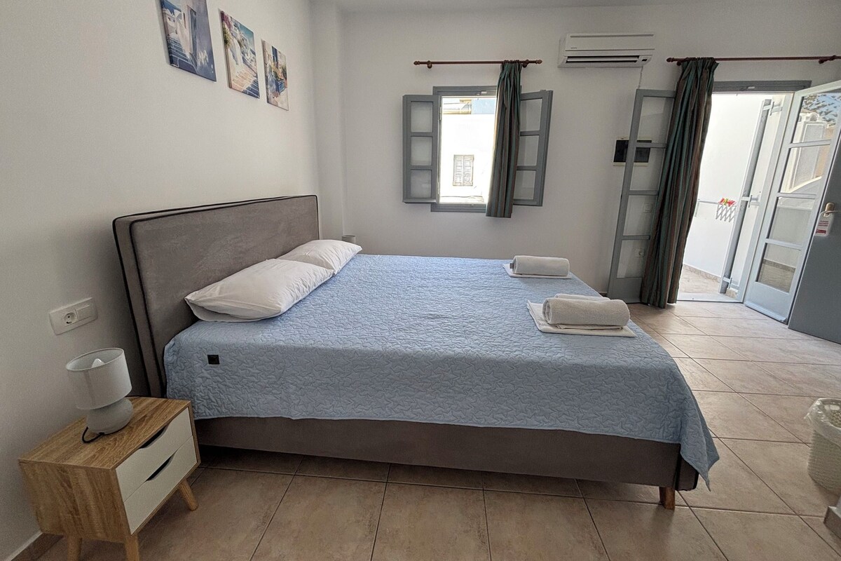 Un dormitorio con cama, mesita de noche, ventana con cortinas y puerta que da a un balcón.
