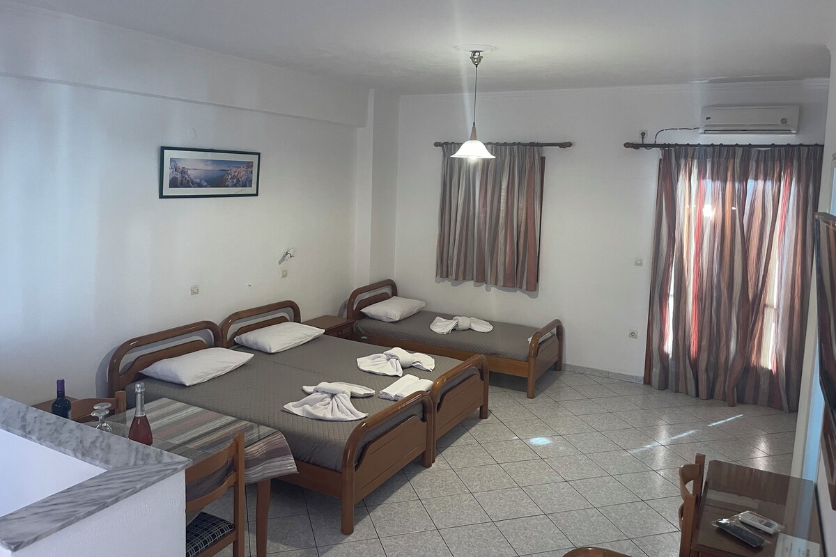 A hotel room with three wooden beds, a small table with chairs, a ceiling light, and an air conditioning unit.