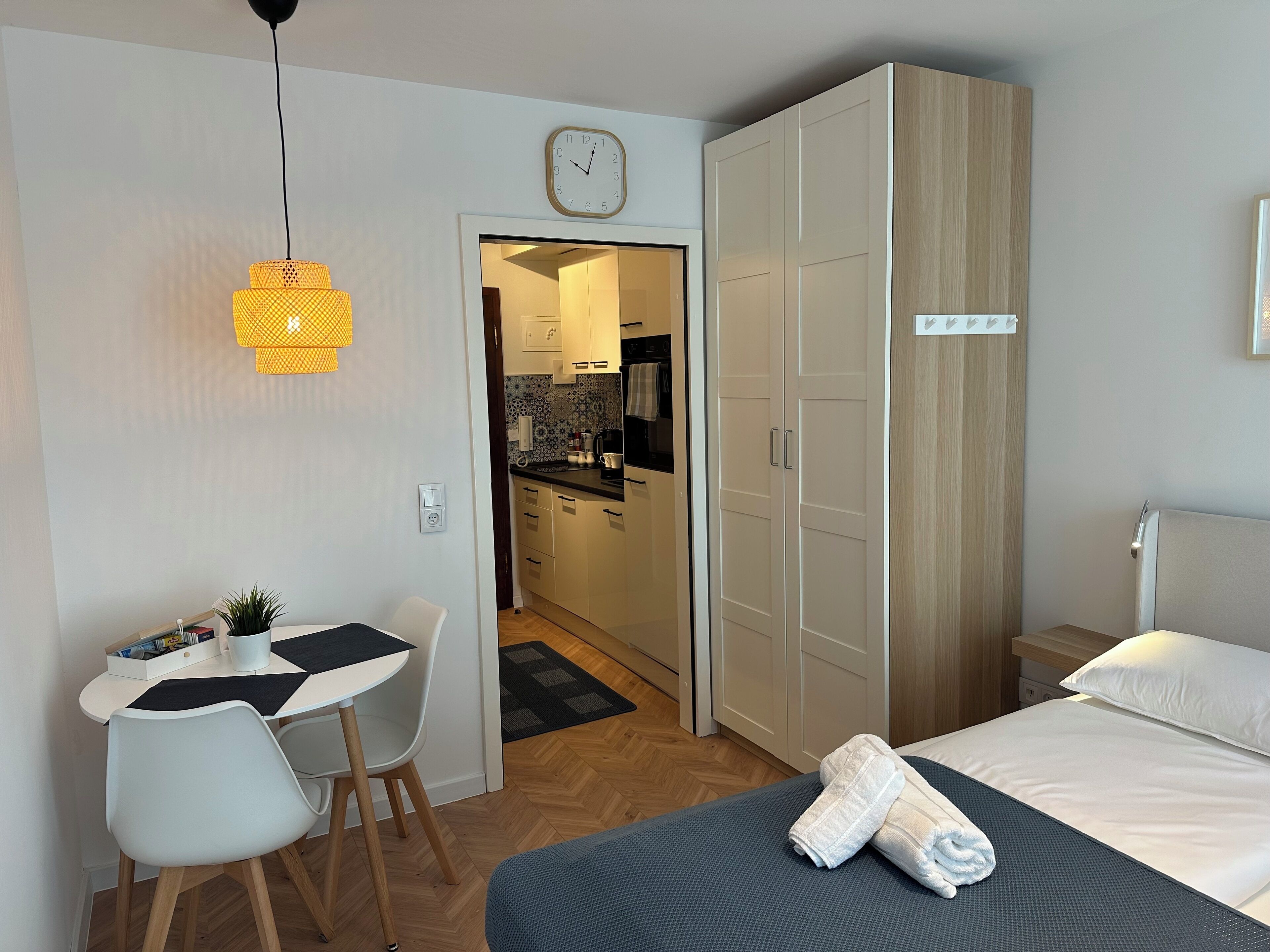 Een moderne hotelkamer met een bed, een bureau met stoel, een kledingkast, en een keukenruimte zichtbaar door een open deur.