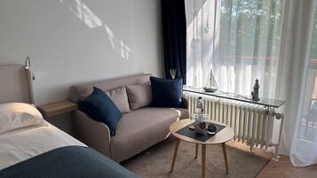 A modern hotel room with a sofa, a small table with a bottle and glasses, a radiator, and a view of the outdoors.