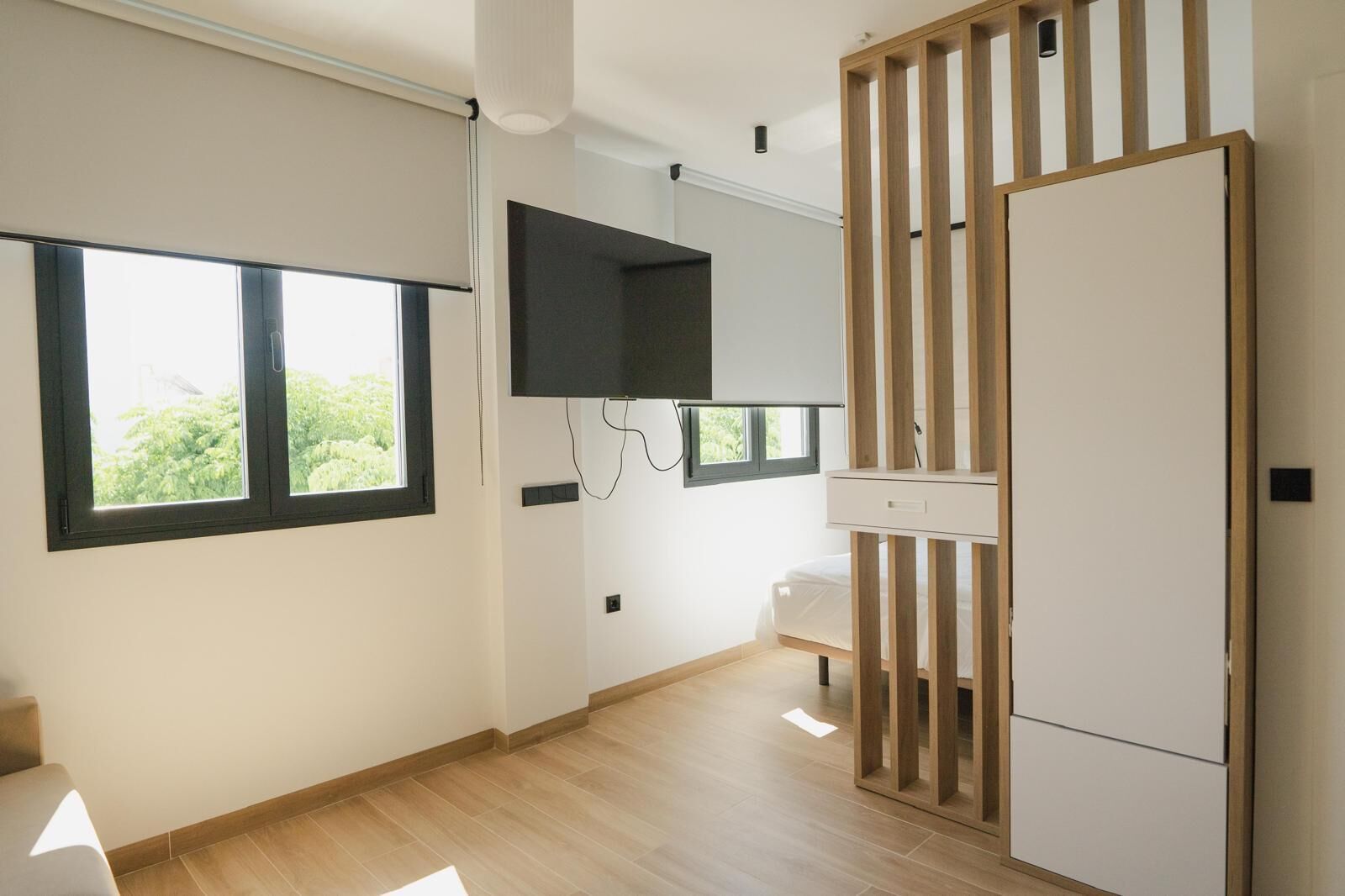 A modern, minimalist room with large windows, a flat-screen TV, and a wooden wardrobe.