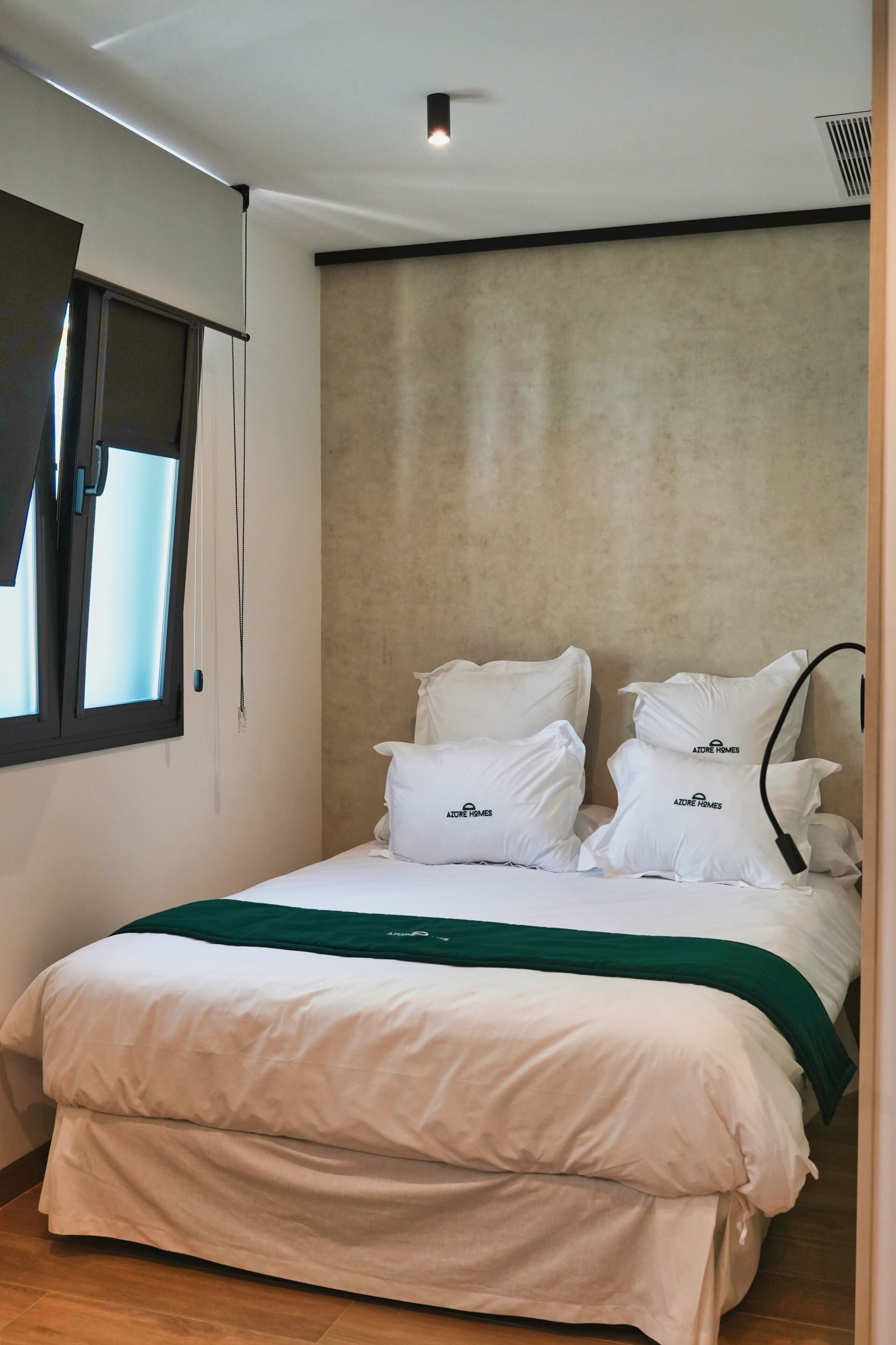 A neatly made bed with white bedding and a green throw, a window with blinds, and a textured beige wall.