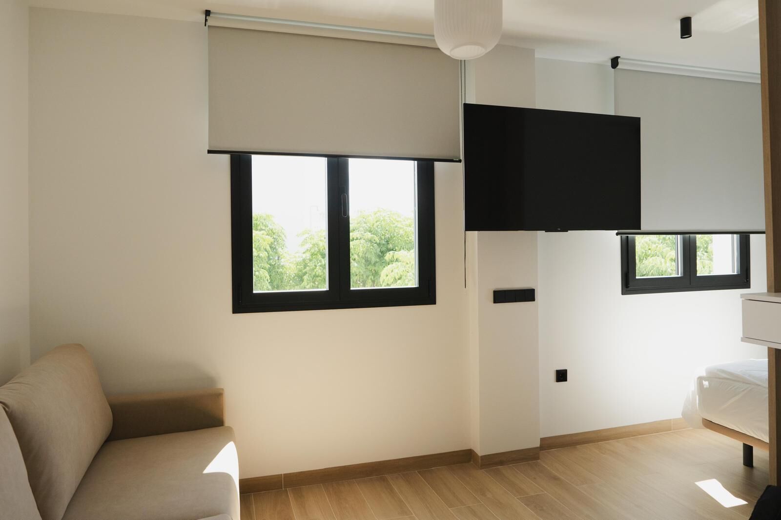 A modern, minimalistic room with a large window, a flat-screen TV mounted on the wall, and a bed with a white mattress.