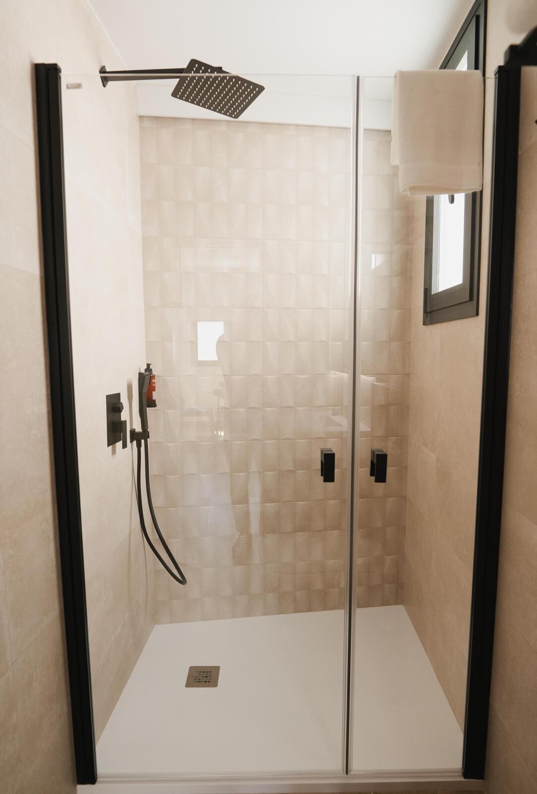 Ducha a pie con mampara de vidrio, azulejos color beige, un cabezal de ducha manual y una ventana pequeña.