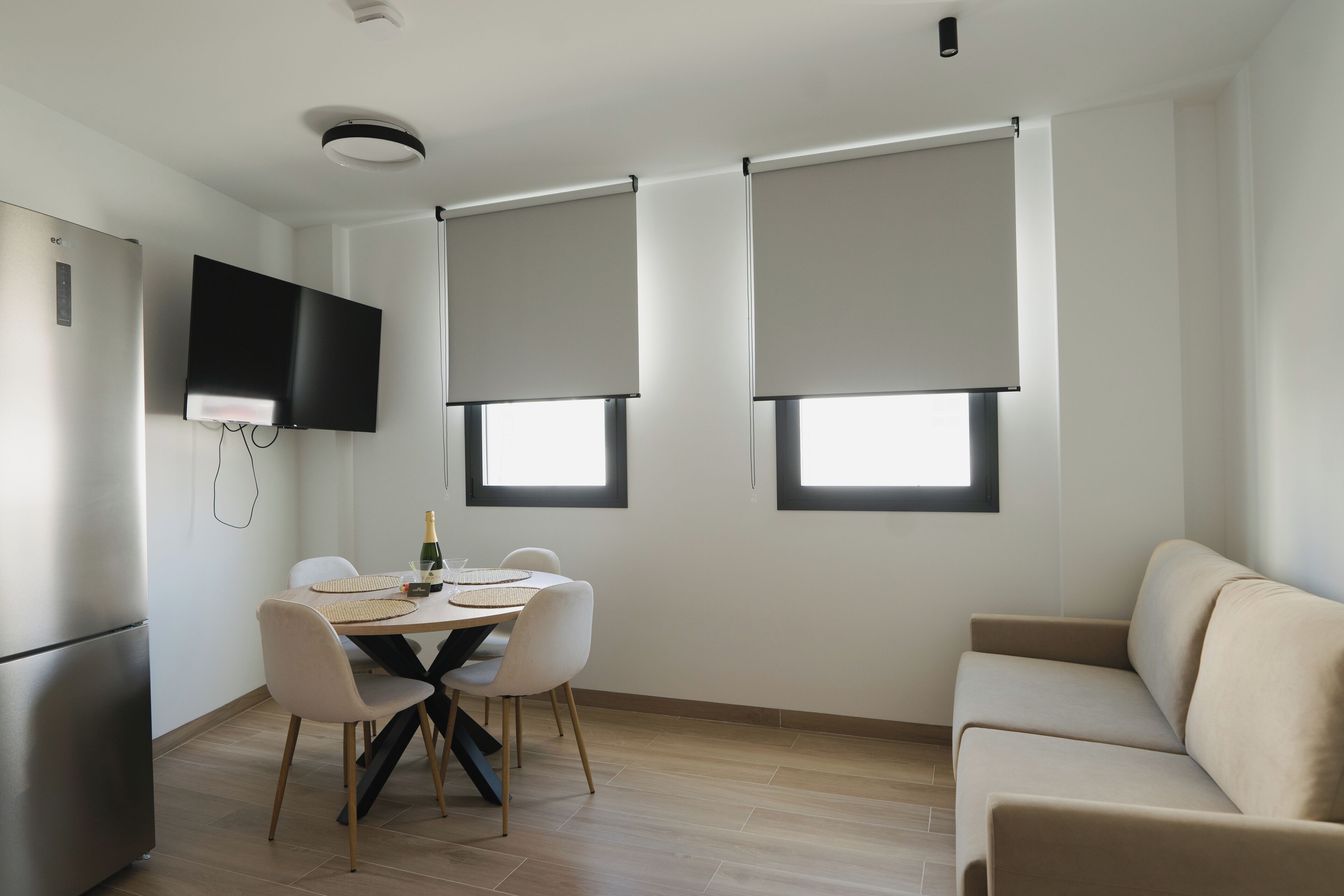 A modern kitchen with a dining area, a flat-screen TV mounted on the wall, and a sofa.