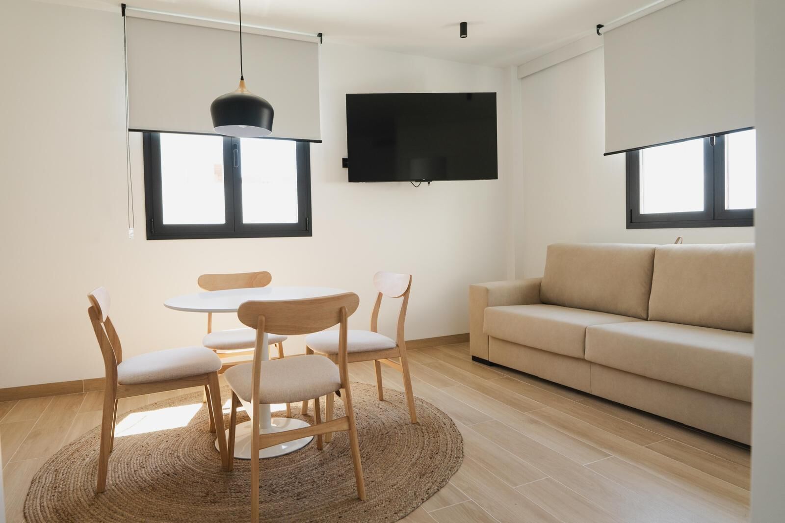 Ein modernes Wohnzimmer mit einem runden Tisch, vier Stühlen, einem beigefarbenen Sofa, einem Flachbildfernseher und einem großen Fenster.