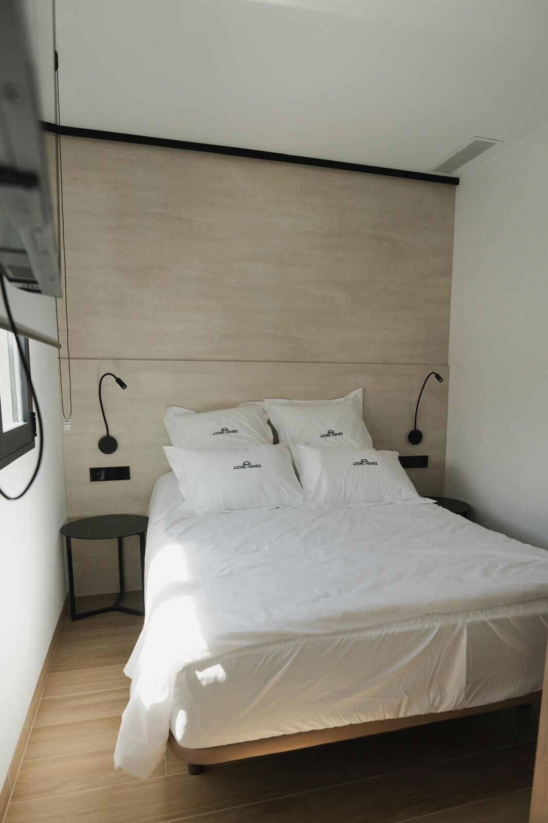 Quarto moderno com cabeceira de madeira, cama com roupa de cama branca, duas mesas de cabeceira e uma luminária de parede.