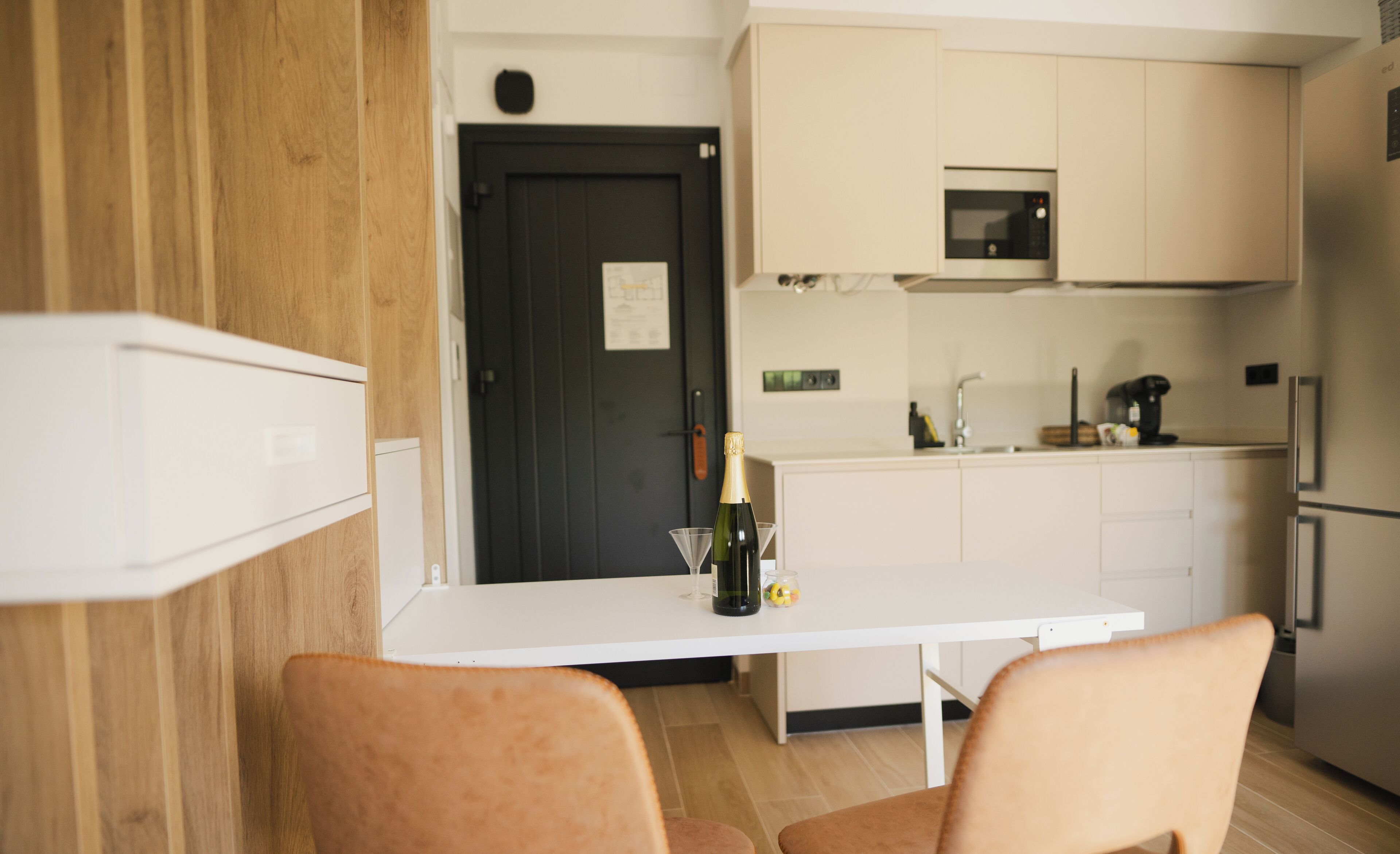 A modern kitchen with white cabinets, a black door, and a microwave. There is a table with chairs and a bottle of champagne.
