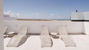 Une terrasse sur le toit, dotée de quatre chaises longues blanches et de petites tables rondes, offrant une vue sur une plage et un ciel dégagé.