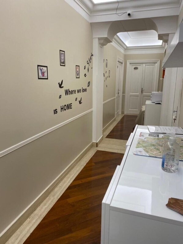 A hallway with a wooden floor, white walls, and a ceiling with recessed lighting. There are framed pictures on the wall and a white table with a map and a bottle on it.