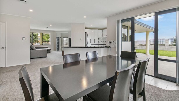 Une cuisine moderne avec des armoires blanches et un grand îlot central, un coin repas avec une table et des chaises aux couleurs sombres, et une porte-fenêtre coulissante donnant sur une terrasse avec vue sur l’extérieur.