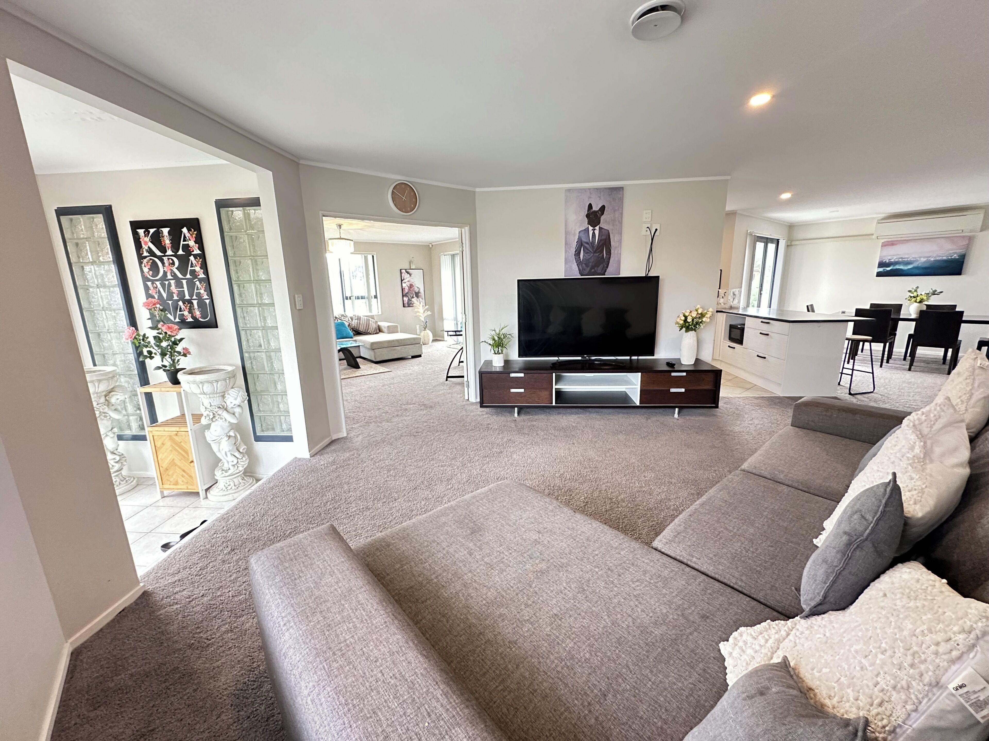 A modern living room with a large flat-screen TV, grey sofas, a dining area with a kitchen in the background, and a hallway leading to another room.
