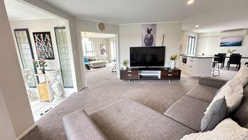 A modern living room with a large flat-screen TV, grey sofas, a dining area with a kitchen in the background, and a hallway leading to another room.