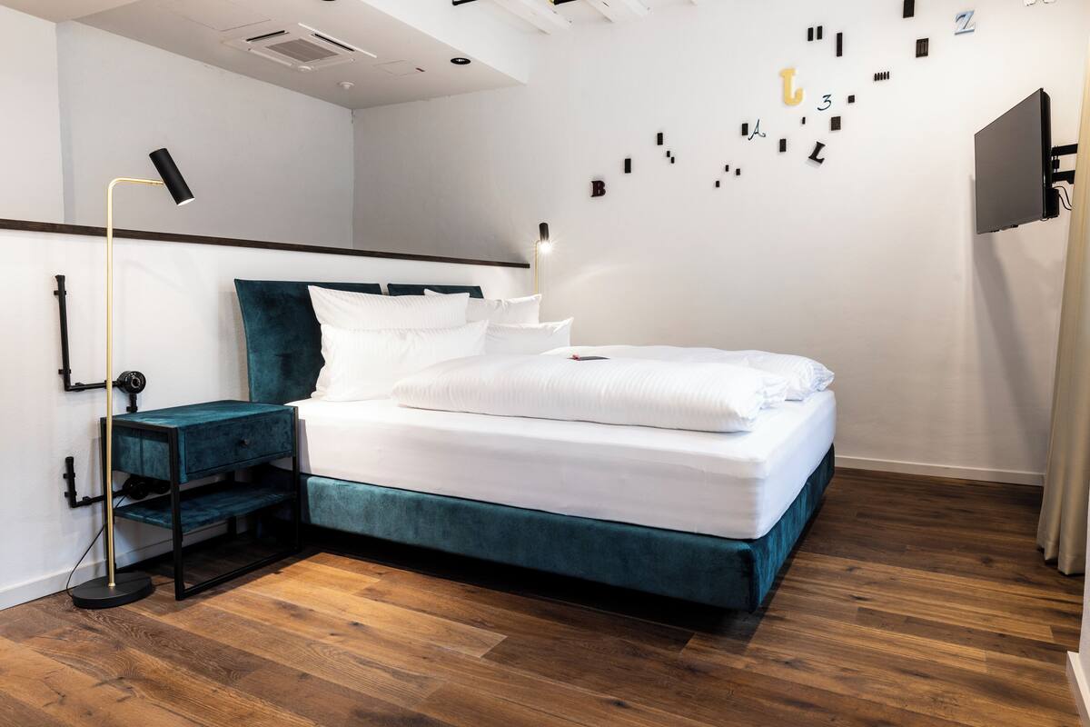 A modern bedroom with a teal upholstered bed, white bedding, a bedside table with a lamp, and a wall-mounted TV.