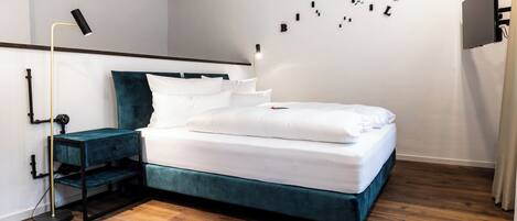 A modern bedroom with a teal upholstered bed, white bedding, a bedside table with a lamp, and a wall-mounted TV.