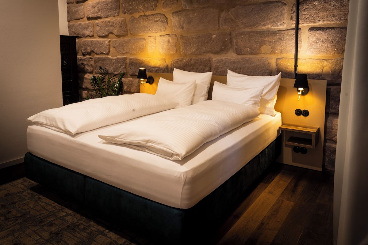 A neatly made bed with white linens, two bedside lamps, and a stone wall behind.