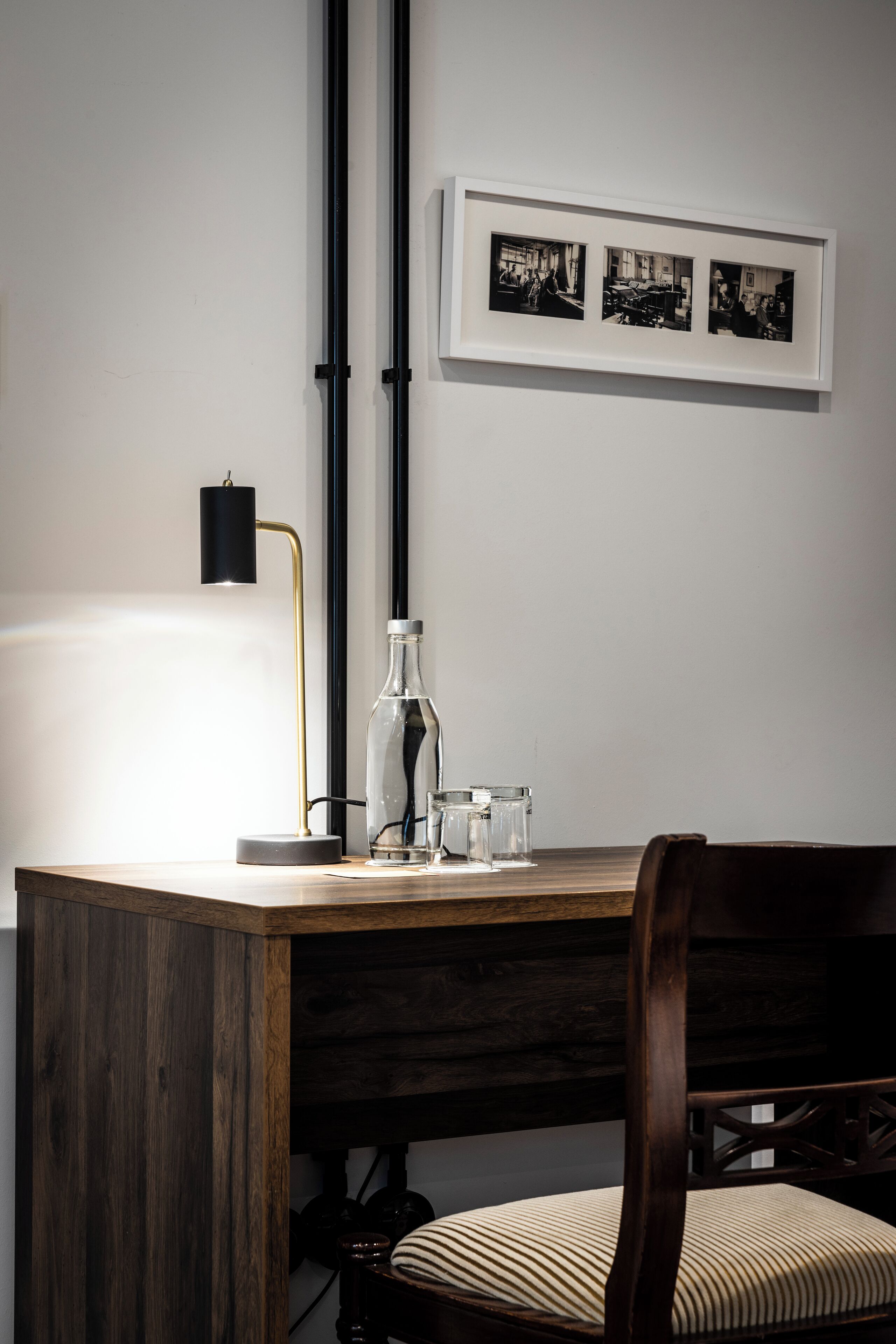 A wooden desk with a lamp, a bottle, and glasses. There is a chair with a striped cushion. A framed photo is on the wall above the desk.