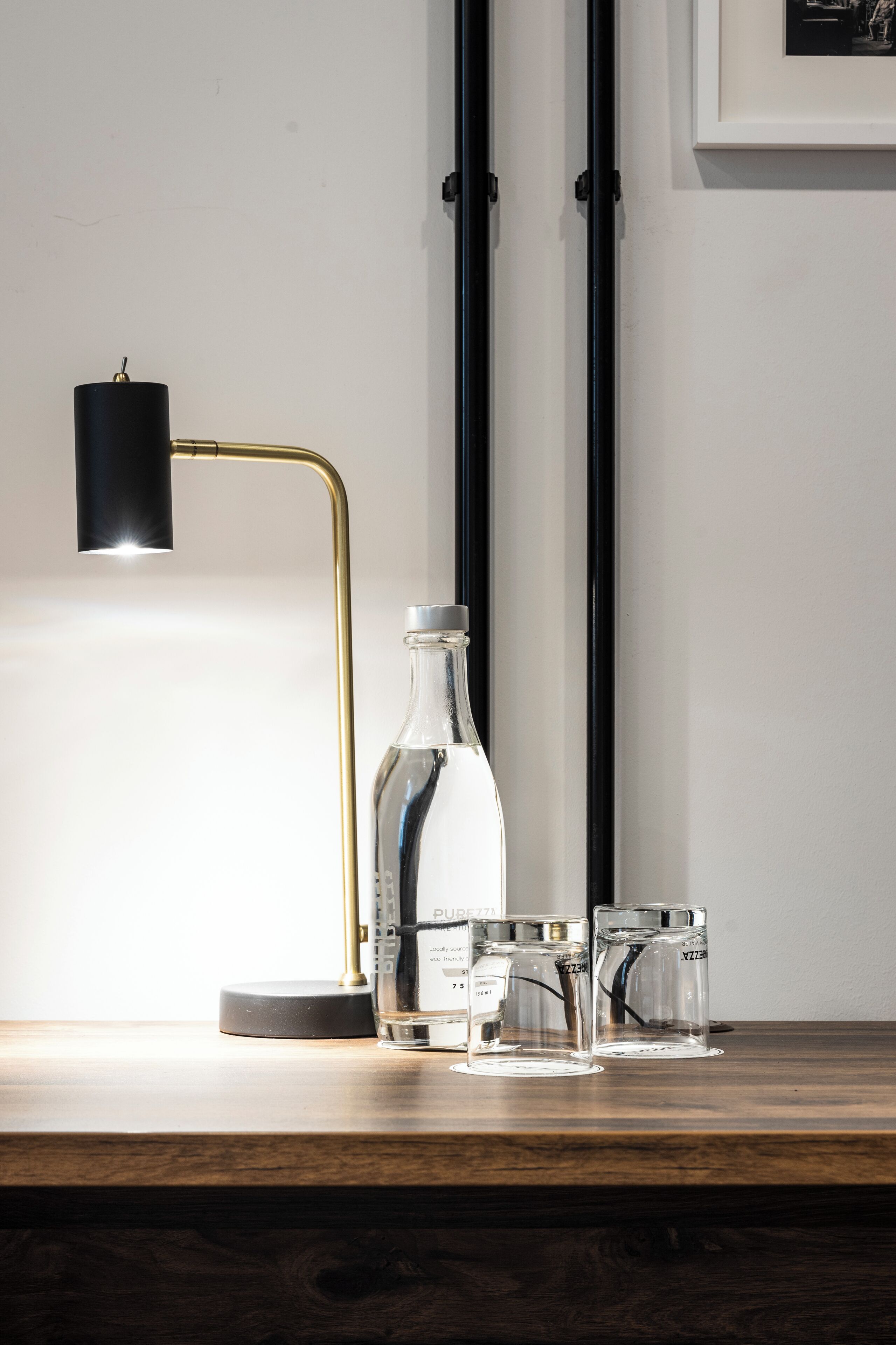 Una mesa de madera con una botella de vidrio transparente, dos vasos de vidrio y una lámpara negra con base dorada.