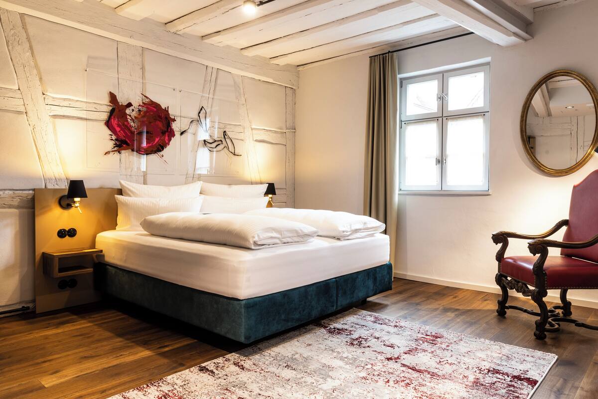 A bedroom with a bed, bedside lamps, a red chair, a window, and a mirror.