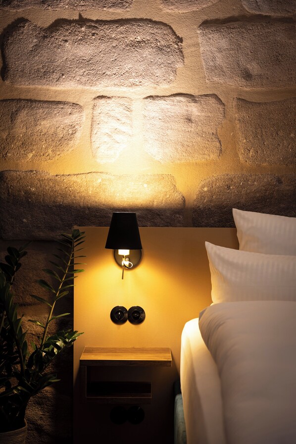 Un dormitorio con pared de piedra, una lámpara de noche, una cama con ropa de cama blanca y una planta en maceta.