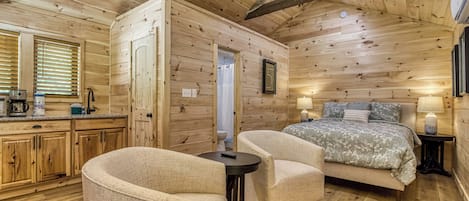 A cozy cabin-style room with a wooden ceiling, a bed with a floral bedspread, two armchairs, a small table, and a kitchenette with a coffee maker.