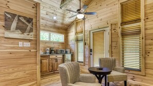 A wooden interior with a ceiling fan, a kitchen area, and two chairs near a window with blinds.