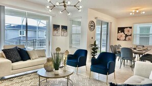 Ein modernes Wohnzimmer mit einer Couch, zwei blauen Sesseln, einem Esstisch mit Stühlen und einem großen Fenster mit Jalousien.
