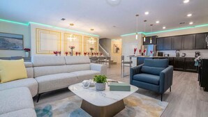 A modern living room with a sofa, armchair, and coffee table, connected to a kitchen area with dark cabinets and a dining area with a staircase. - Epic 10BR Star Wars Villa • Theater • Pool (Davenport)