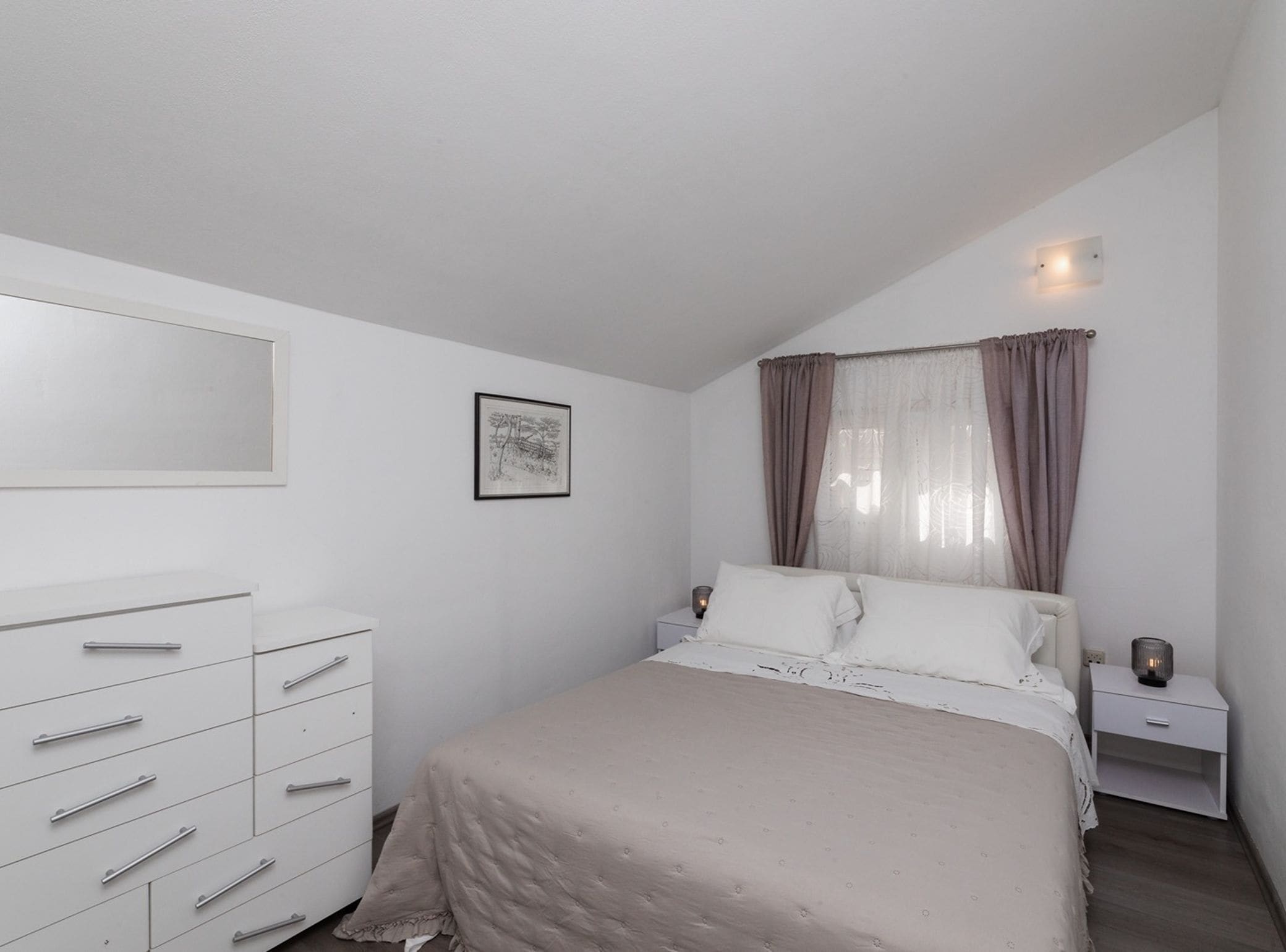 Une chambre à coucher comprenant un lit, une commode, une table de chevet, une lampe fixée au mur et un tableau encadré.