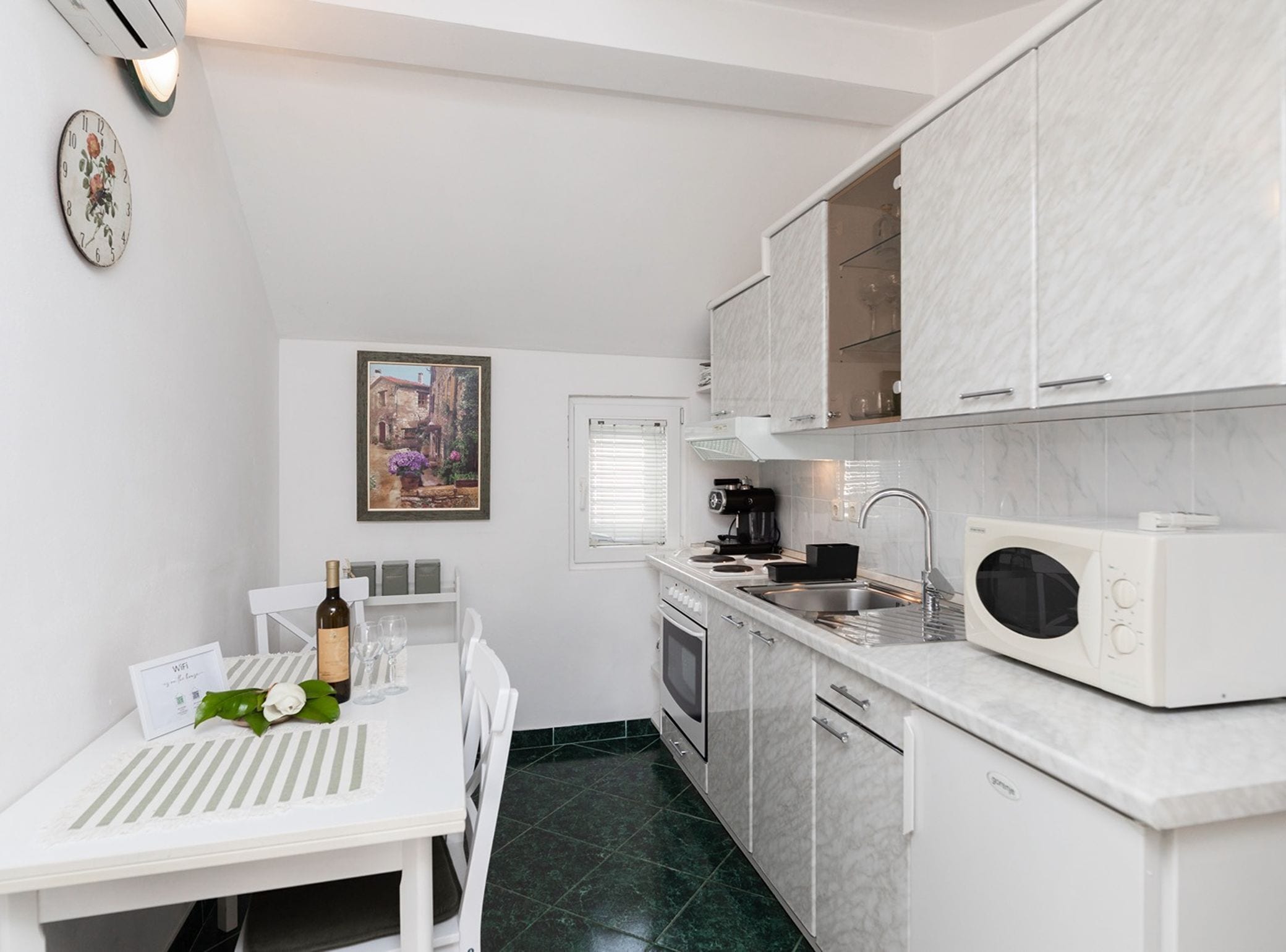 A compact kitchen with white cabinetry, a microwave, a coffee maker, and a dining table with a rose.