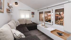 A modern living room with a dark brown sofa, white walls, a wooden side table, and a window with a view of buildings. - Apartments Roda - (Supetar)