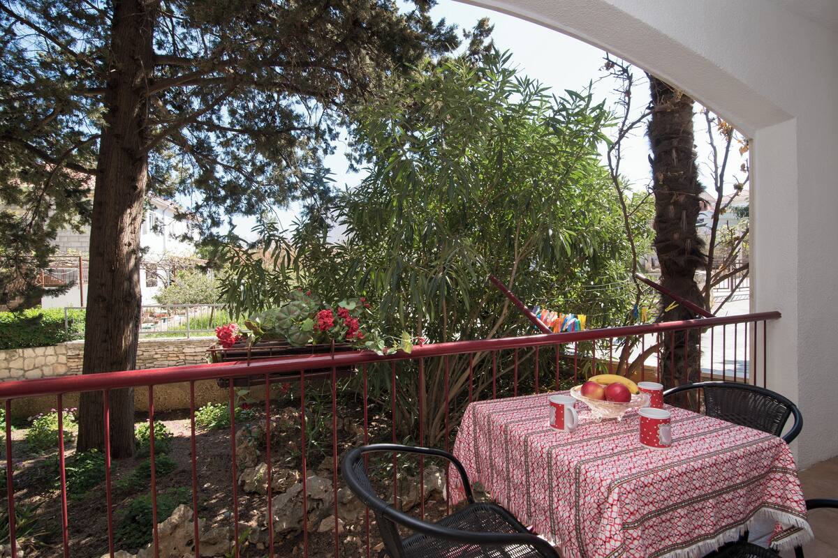 A balcony with a table set for two, surrounded by greenery and a red railing.