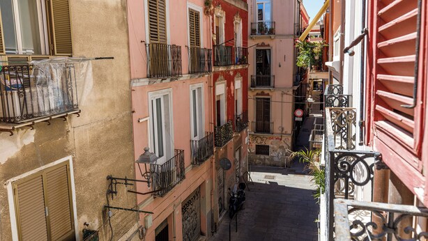 Une rue étroite, bordée de bâtiments de chaque côté, avec des balcons dotés de garde-corps en métal et de volets.