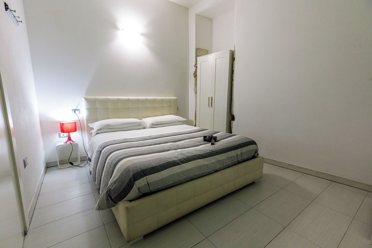Ein Schlafzimmer mit einem Bett, einem Nachttisch mit Lampe, einem Kleiderschrank und einem Spiegel an der Wand.